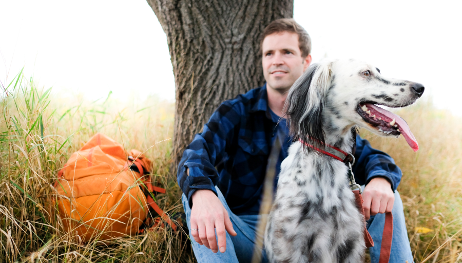 Mężczyzna siedział przed drzewem z pomarańczowym plecakiem na trawie, a jego pies siedział przed nim na smyczy